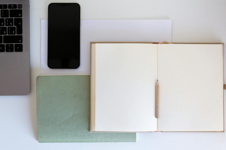 Top view of a minimalist workspace featuring an open notebook, pencil, laptop, and smartphone on a white desk.
